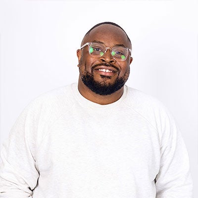 Tumi Sotire smiles at the camera. He has short, black hair and a beard. He wears glasses and a white jumper. 